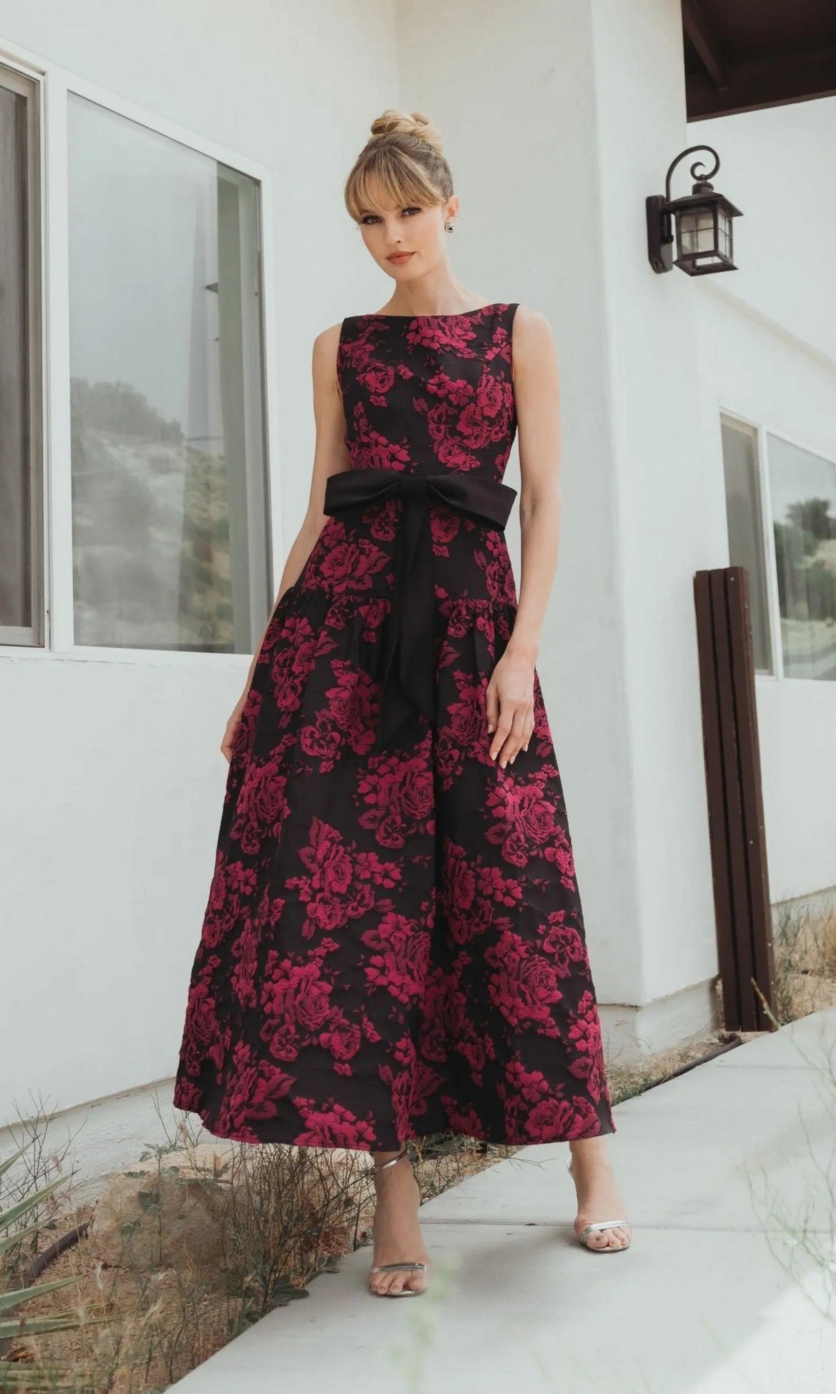A woman in a Mac Duggal 37091 Floral Drop-Waist Party Dress in Magenta Multi with a black sash stands outside a modern white building.